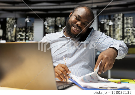 Black young businessman takes phone call for a remote chat with expert, joining a collaborative discussion on telephone line to set the agenda. Adaptability and attentiveness after hours. Black young businessman takes phone call for a remote chat with expert, joining a collaborative discussion on telephone line to set the agenda. Adaptability and attentiveness after hours. 138022133