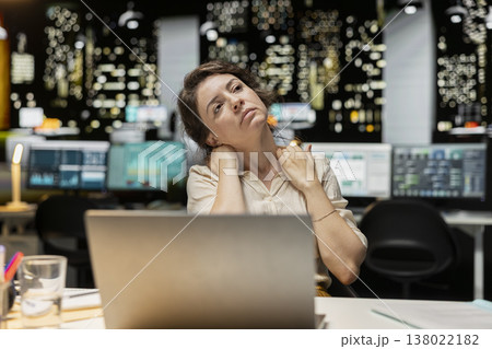 Office employee stretching her upper back and neck muscles, correcting the posture and neck stiffness for relief and relaxation. Intense office activity due to overtime late at night. 138022182