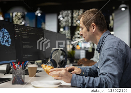 Admin eating croissant while using computer in tech startup office to develop programs with AI. IT professional enjoying snack break while debugging applications with artificial intelligence Admin eating croissant while using computer in tech startup office to develop programs with AI. IT professional enjoying snack break while debugging applications with artificial intelligence 138022193