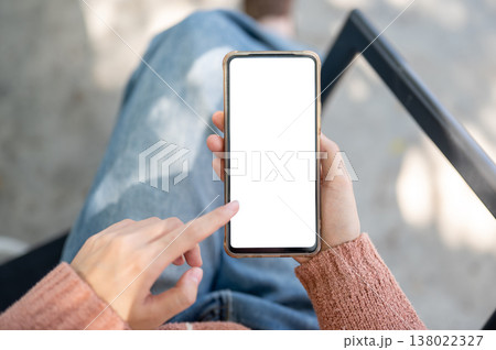 A woman's hand holding white screen phone with finger pointing sitting crossed legs on outdoor bench A woman's hand holding white screen phone with finger pointing sitting crossed legs on outdoor bench 138022327