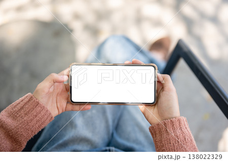 A woman's hands holding white screen phone horizontally sitting on outdoor bench over concrete floor 138022329