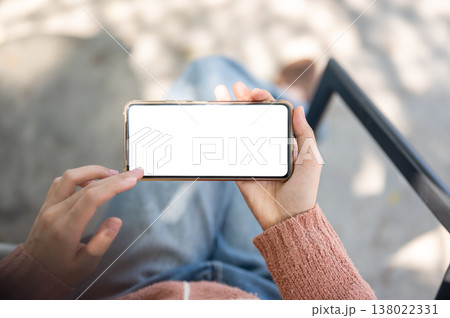 A woman's hand holding white screen phone horizontally sitting on outdoor bench over concrete floor. A woman's hand holding white screen phone horizontally sitting on outdoor bench over concrete floor. 138022331