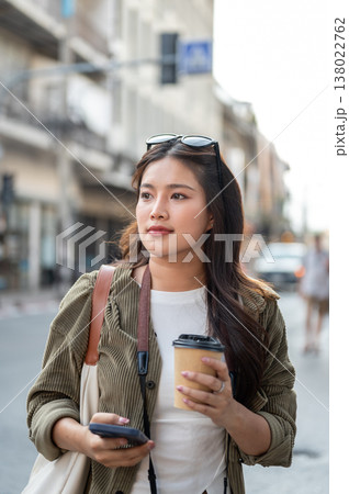 Pretty asian woman holding coffee and phone while standing or walking by road street in local town. Pretty asian woman holding coffee and phone while standing or walking by road street in local town. 138022762