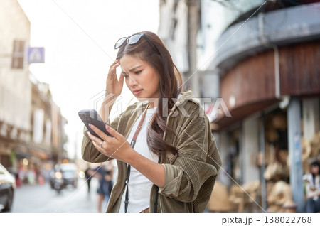 Confused asian woman scratching head looking at phone while standing by road street in local town. Confused asian woman scratching head looking at phone while standing by road street in local town. 138022768