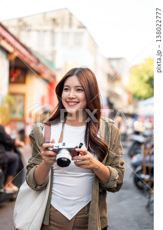 Pretty asian woman holding camera looking ahead standing or walking in local street market of town. 138022777