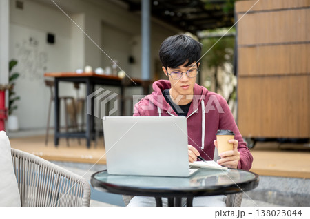 Glasses asian man student freelancer holding coffee as writing in book aside laptop at table in cafe Glasses asian man student freelancer holding coffee as writing in book aside laptop at table in cafe 138023044