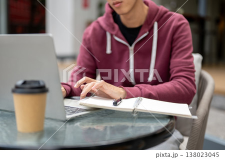 Close up of asian man student freelancer typing on laptop aside notebook and coffee at table in cafe Close up of asian man student freelancer typing on laptop aside notebook and coffee at table in cafe 138023045