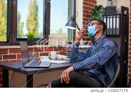 African american manager seated in brick wall office looking pensive and tapping his forehead with a pen. Businessman with face mask, gazing outside the window and thinking deeply on company projects. African american manager seated in brick wall office looking pensive and tapping his forehead with a pen. Businessman with face mask, gazing outside the window and thinking deeply on company projects. 138027179