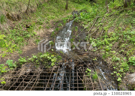 Small Forest Waterfall and Stream Flowing Over a Metal Grate on a Green Spring Hillside 138028965