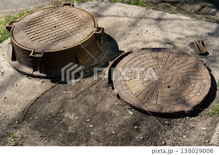 Rusty Open Manhole Cover and Raised Sewer Lid on Cracked Asphalt Sidewalk Surface Rusty Open Manhole Cover and Raised Sewer Lid on Cracked Asphalt Sidewalk Surface 138029006