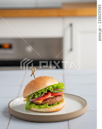 Meat burger with tomato, cheese, and lettuce on a plate on a table against a white kitchen Meat burger with tomato, cheese, and lettuce on a plate on a table against a white kitchen 138029365