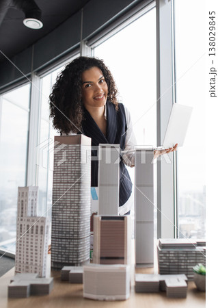 Female architect presenting a scale skyscraper model by a floor-to-ceiling window while using a laptop 138029845