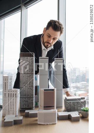 A focused architect in a suit examines a collection of miniature skyscrapers on a table, contemplating urban development A focused architect in a suit examines a collection of miniature skyscrapers on a table, contemplating urban development 138029872
