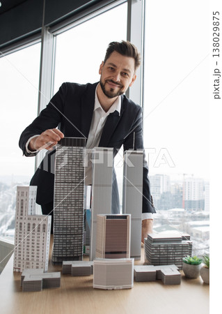 Smiling architect in a suit examines a detailed scale model of high-rise buildings on a table by a large office window overlooking the city Smiling architect in a suit examines a detailed scale model of high-rise buildings on a table by a large office window overlooking the city 138029875