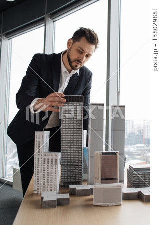 A focused architect in a suit examines a detailed scale model of a city skyline, considering urban development and design 138029881