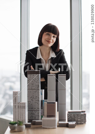 A professional woman stands behind a collection of miniature city buildings, suggesting urban planning or real estate development A professional woman stands behind a collection of miniature city buildings, suggesting urban planning or real estate development 138029900