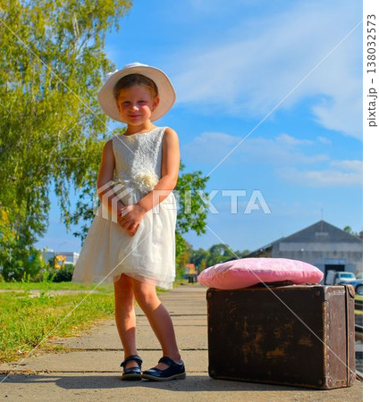 A cute little girl in a white dress and hat is waiting for her vacation. The little girl is waiting for the vacation train to arrive. The concept of a carefree childhood, summer vacation, and family. A cute little girl in a white dress and hat is waiting for her vacation. The little girl is waiting for the vacation train to arrive. The concept of a carefree childhood, summer vacation, and family. 138032573