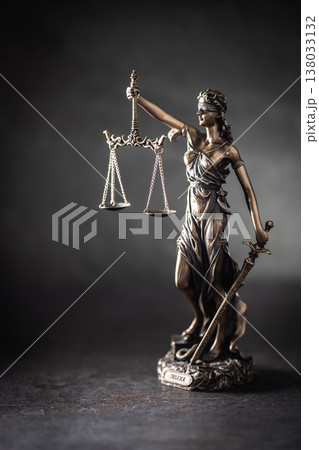 A bronze statue of Justice stands against a dark, textured background with dramatic side lighting. The figure holds scales symbolizing justice in one hand and a sword in the other, her eyes covered by A bronze statue of Justice stands against a dark, textured background with dramatic side lighting. The figure holds scales symbolizing justice in one hand and a sword in the other, her eyes covered by 138033132