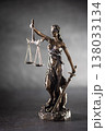 A bronze statue of Justice stands against a dark, textured background with dramatic side lighting. The figure holds scales symbolizing justice in one hand and a sword in the other, her eyes covered by 138033134