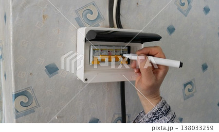A hand holding a black marker applies labels to the circuit breakers in a residential distribution board, and also marks the meter mounted on the wall. The breakers for various electrical circuits A hand holding a black marker applies labels to the circuit breakers in a residential distribution board, and also marks the meter mounted on the wall. The breakers for various electrical circuits 138033259
