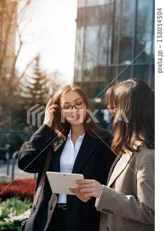 Women discussing business ideas while using a tablet in an outdoor office setting near city buildings during daylight hours Women discussing business ideas while using a tablet in an outdoor office setting near city buildings during daylight hours 138034504