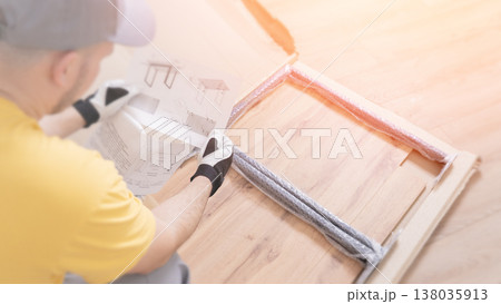 A middle-aged Caucasian man is assembling furniture on the floor using a screwdriver, meticulously fastening wooden components, surrounded by tools and assembly instructions in a contemporary domestic 138035913
