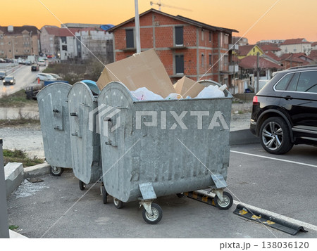 Overflowing metal garbage containers on a neighborhood street at sunset near residential buildings. Waste collection, urban sanitation, municipal service, recycling challenge, everyday infrastructure Overflowing metal garbage containers on a neighborhood street at sunset near residential buildings. Waste collection, urban sanitation, municipal service, recycling challenge, everyday infrastructure 138036120