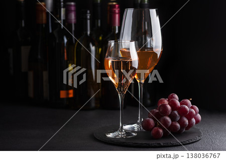 TWo  glasses of white and sparkling rose wine with bunch of grapes on dark slate surface against bottles in background 138036767
