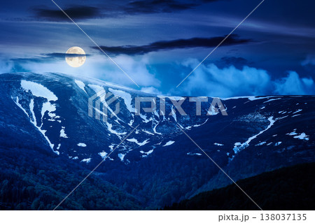 mysterious nature image of ridge with snow capped tops in spring landscape. mountain background wallpaper at night. full moon over greenery scenery with forested slopes under sky with clouds 138037135