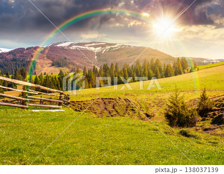 sunset over mountainous countryside landscape in spring. wooden fence and grassy meadow on a green hill. distant mountain ridge with snow capped tops. cloudy sky and rainbow 138037139