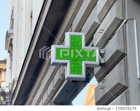 Green illuminated pharmacy cross sign mounted on a stone building facade in the city center. Healthcare wayfinding, medical retail identity, and urban signage in a historic streetscape context 138039274