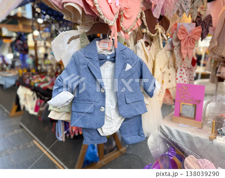 Baby boy formal outfit with blazer, shorts, and bow tie hanging at a boutique stall. Infant occasion wear, ceremonial styling, gift buying, and elegant retail presentation for christening or 138039290