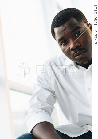 Young man sits indoors, looking at the camera in a bright setting 138041118
