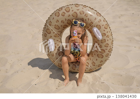 A girl in a swimsuit is sitting on the beach in an inflatable circle holding a cupcake with candles in the shape of the number five, thinking about a wish A girl in a swimsuit is sitting on the beach in an inflatable circle holding a cupcake with candles in the shape of the number five, thinking about a wish 138041430