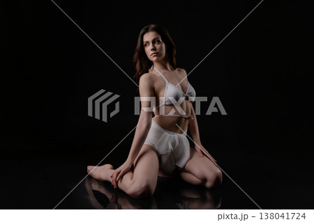 Pensive brunette woman in white bikini kneeling in water on a black studio background. 138041724
