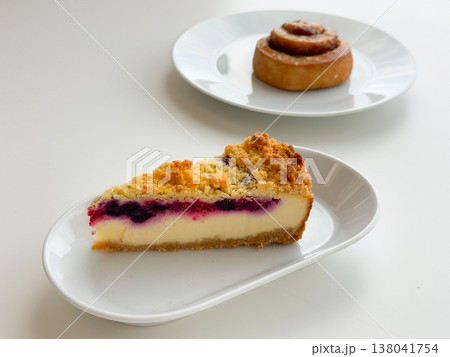 Blueberry cheesecake slice with cinnamon roll in soft focus on white tableware. Bakery assortment styling, dessert pairing, cafe selection, pastry presentation, minimal food composition concept Blueberry cheesecake slice with cinnamon roll in soft focus on white tableware. Bakery assortment styling, dessert pairing, cafe selection, pastry presentation, minimal food composition concept 138041754