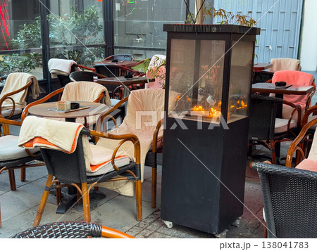 Outdoor restaurant terrace with heater and blankets under large umbrella in cool weather. Seasonal comfort strategy, customer retention, hospitality adaptation, energy usage, urban dining environment Outdoor restaurant terrace with heater and blankets under large umbrella in cool weather. Seasonal comfort strategy, customer retention, hospitality adaptation, energy usage, urban dining environment 138041783