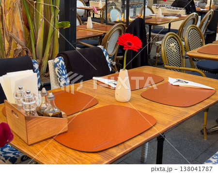 Restaurant table setting with Gerbera jamesonii flower in vase and condiment set on wooden surface. Dining preparation, service organization, hospitality detail, customer readiness, table arrangement 138041787