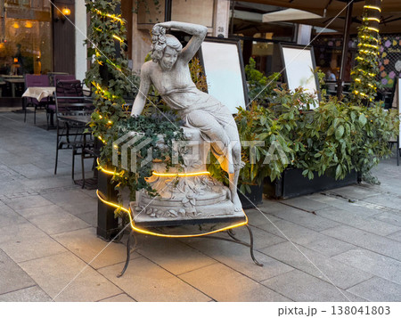 Classical stone sculpture of reclining woman decorated with ivy and light garland near cafe entrance. Artistic decoration, urban aesthetics, cultural reference, hospitality branding, exterior styling 138041803