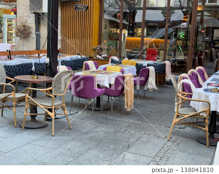 Outdoor restaurant tables with purple chairs and blankets arranged on city terrace. Seasonal seating comfort, textile layering, hospitality service design, customer experience, urban dining 138041810