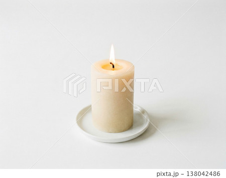 Lit white pillar candle on a small saucer, glowing gently on a white background, home decor Lit white pillar candle on a small saucer, glowing gently on a white background, home decor 138042486