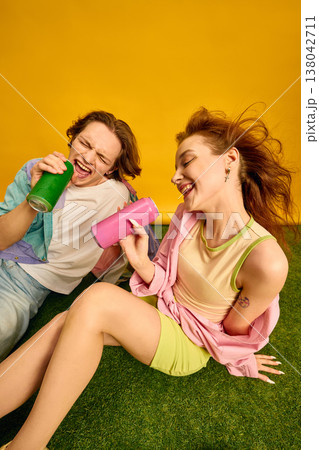 Young couple sitting on grass singing and drinking canned beverages in studio. Young couple sitting on grass singing and drinking canned beverages in studio. 138042711