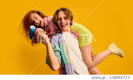 Young man carrying smiling woman on shoulder holding drink cans on bright yellow background. Young man carrying smiling woman on shoulder holding drink cans on bright yellow background. 138042713