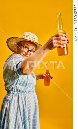 Elderly woman raising bottled drink toward camera making toast on yellow background. 138042729