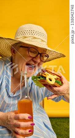 Elderly woman biting burger while holding glass bottle in bright yellow summer scene. Elderly woman biting burger while holding glass bottle in bright yellow summer scene. 138042748