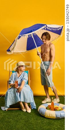 Young man holding beach umbrella over seated elderly woman with bottle in summer setting. Young man holding beach umbrella over seated elderly woman with bottle in summer setting. 138042749