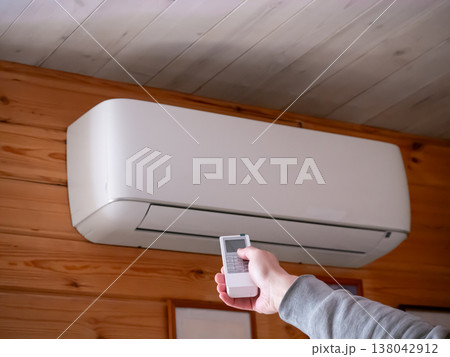 Man holding remote control to switch on air conditioner wall unit. Indoor climate control in living room. Modern comfort technology, cooling system and temperature regulation at home. 138042912