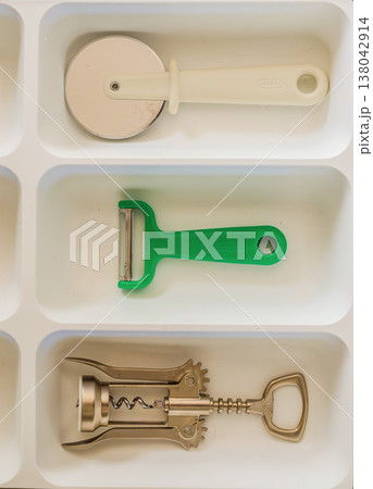 Essential kitchen gadgets: pizza cutter, peeler, and corkscrew neatly organized in a white drawer tray. Essential kitchen gadgets: pizza cutter, peeler, and corkscrew neatly organized in a white drawer tray. 138042914