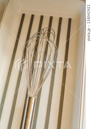 A stainless steel kitchen whisk lies in a modern, minimalist white drawer with a striped bottom. Close-up of a cooking utensil. A stainless steel kitchen whisk lies in a modern, minimalist white drawer with a striped bottom. Close-up of a cooking utensil. 138042921