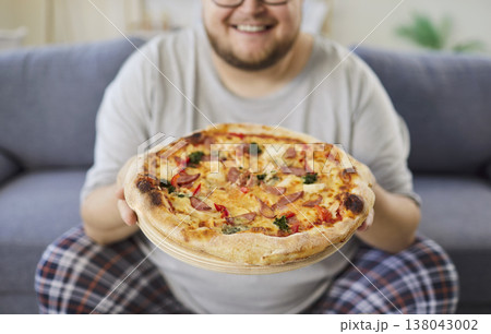Happy man at home presenting pizza on sofa, casual pajamas, comfort moment 138043002